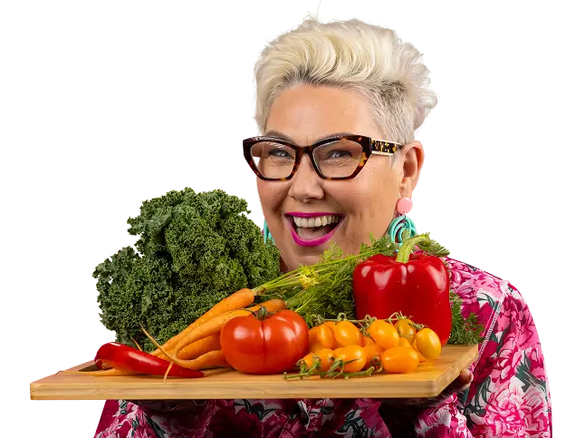 Steph de Sousa smiling while holding a wooden board piled with fresh vegetables, cut out on a transparent background.